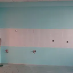 Ściana w trakcie przygotowywania do wykończenia w pomieszczeniu z turkusowymi ścianami, rzędem białych płytek i wystającymi instalacjami hydraulicznymi oraz elektrycznymi, widoczna miotła.