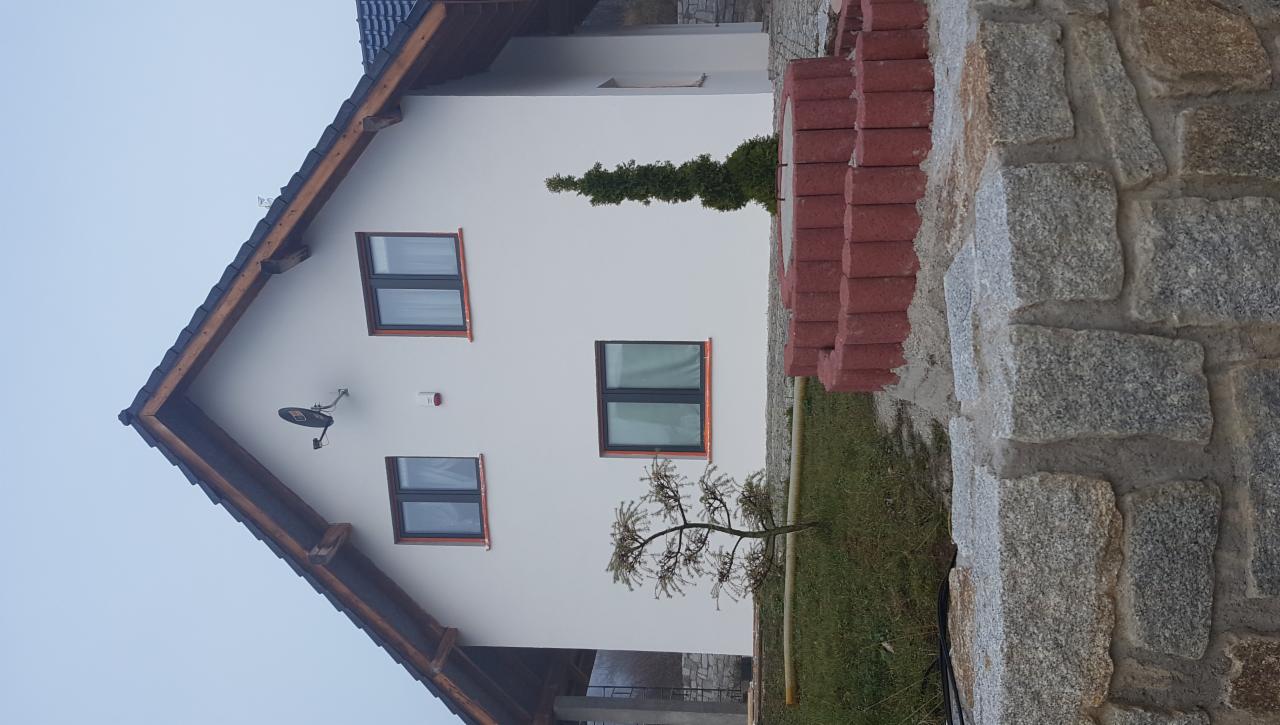 Elewacja domu jednorodzinnego z białym tynkiem, trzema oknami z ciemnymi ramami i anteną satelitarną, widoczna kamienna podmurówka i stos czerwonej kostki brukowej.