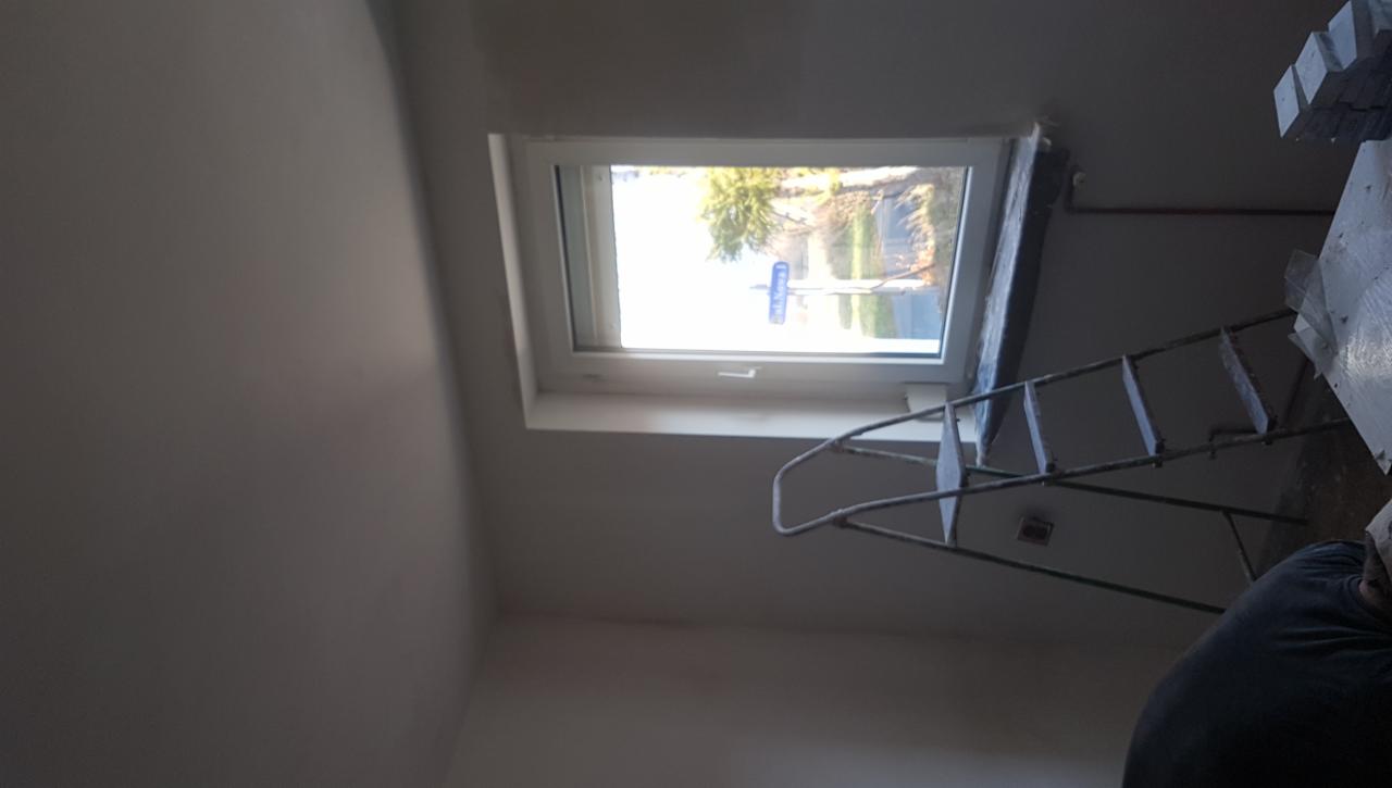 Pokój w trakcie prac wykończeniowych: ściany przygotowane pod malowanie, widoczna drabina i okno dachowe z widokiem na ulicę.