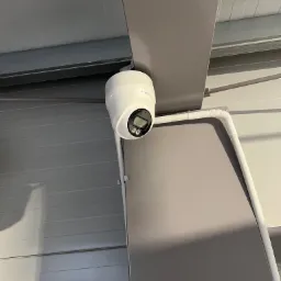 Kamera monitoringu kopułkowa, biała, zamontowana pod sufitem w hali. Widoczne okablowanie w białej listwie. Niewielkie logo producenta na obudowie kamery.