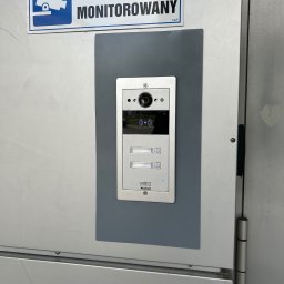 Queter - Zamontowany wideofon Akuvox na szarej podkładce, z tabliczką 'Obiekt Monitorowany' w Słupnie. Widok z bliska na urządzenie przy drzwiach.