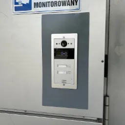 Zamontowany wideofon Akuvox na szarej podkładce, z tabliczką 'Obiekt Monitorowany' w Słupnie. Widok z bliska na urządzenie przy drzwiach.