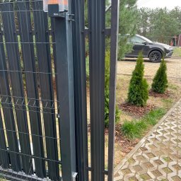 Queter - Automatyczna brama wjazdowa z lampą ostrzegawczą NICE, częściowo otwarta. W tle widoczny samochód osobowy i zieleń. Brama w kolorze grafitowym.