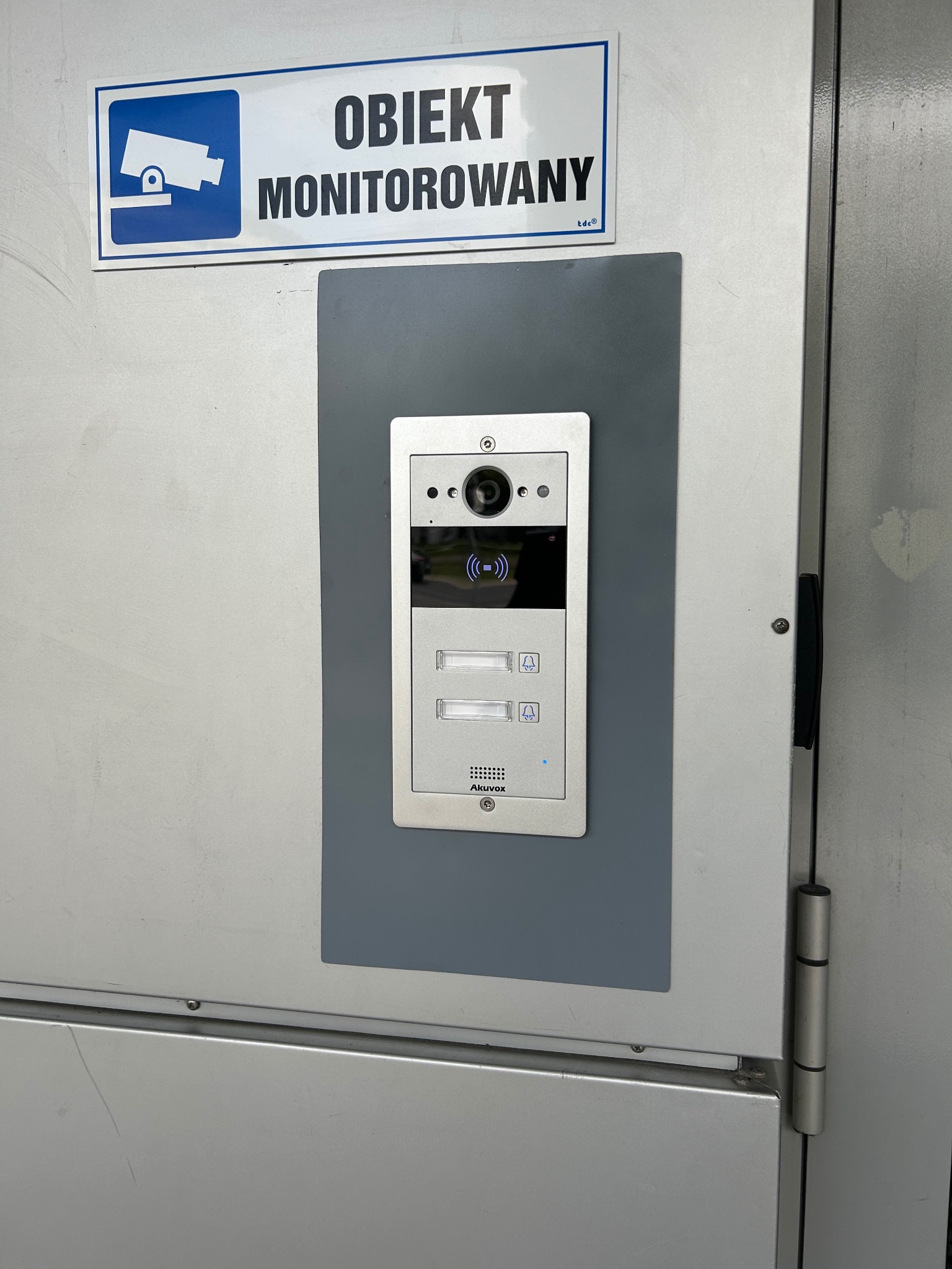 Zamontowany wideofon Akuvox na szarej podkładce, z tabliczką 'Obiekt Monitorowany' w Słupnie. Widok z bliska na urządzenie przy drzwiach.