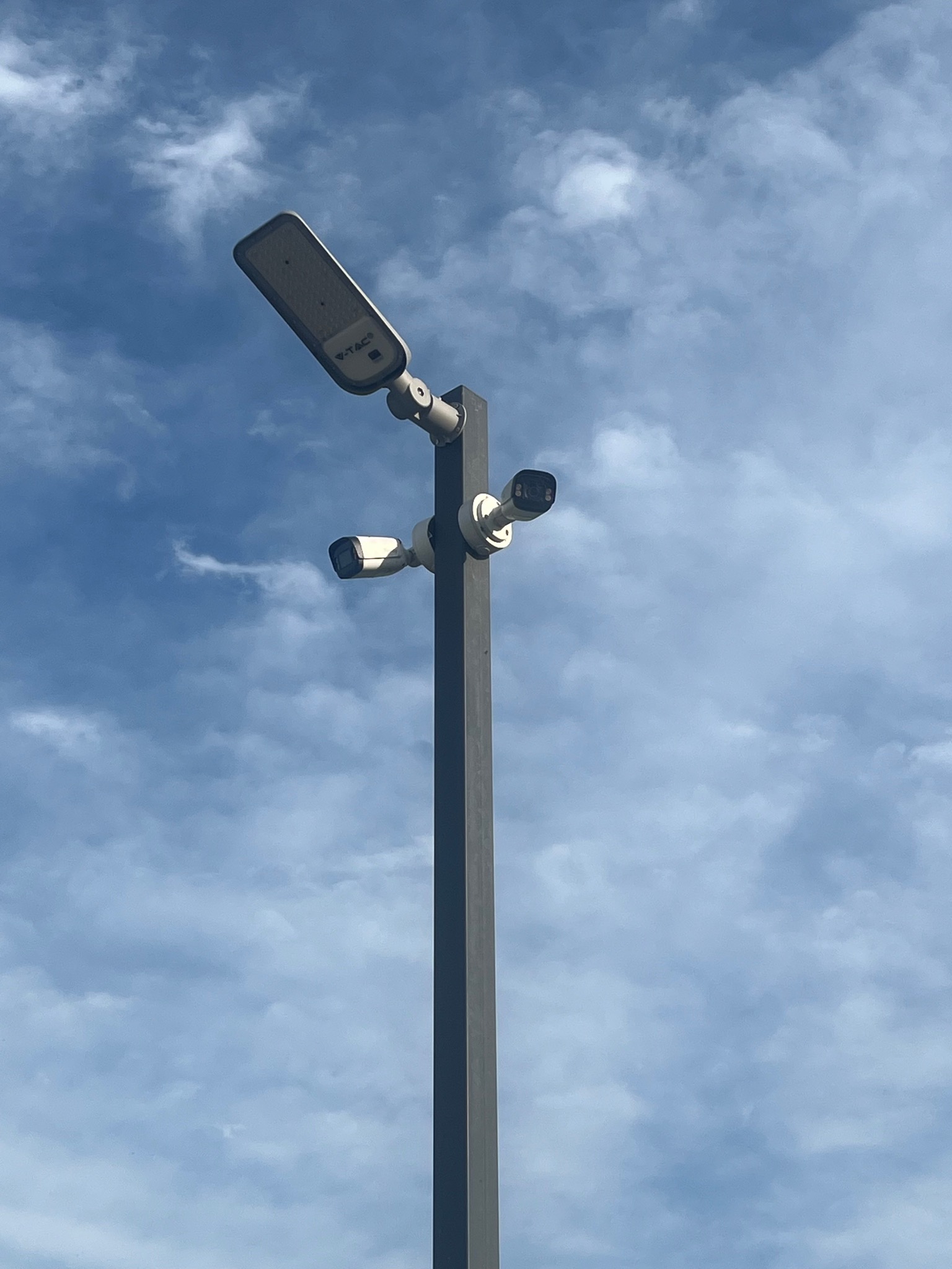Słup oświetleniowy z dwiema kamerami monitoringu na tle błękitnego, zachmurzonego nieba. Kamery skierowane w różne strony. Widok z dołu.