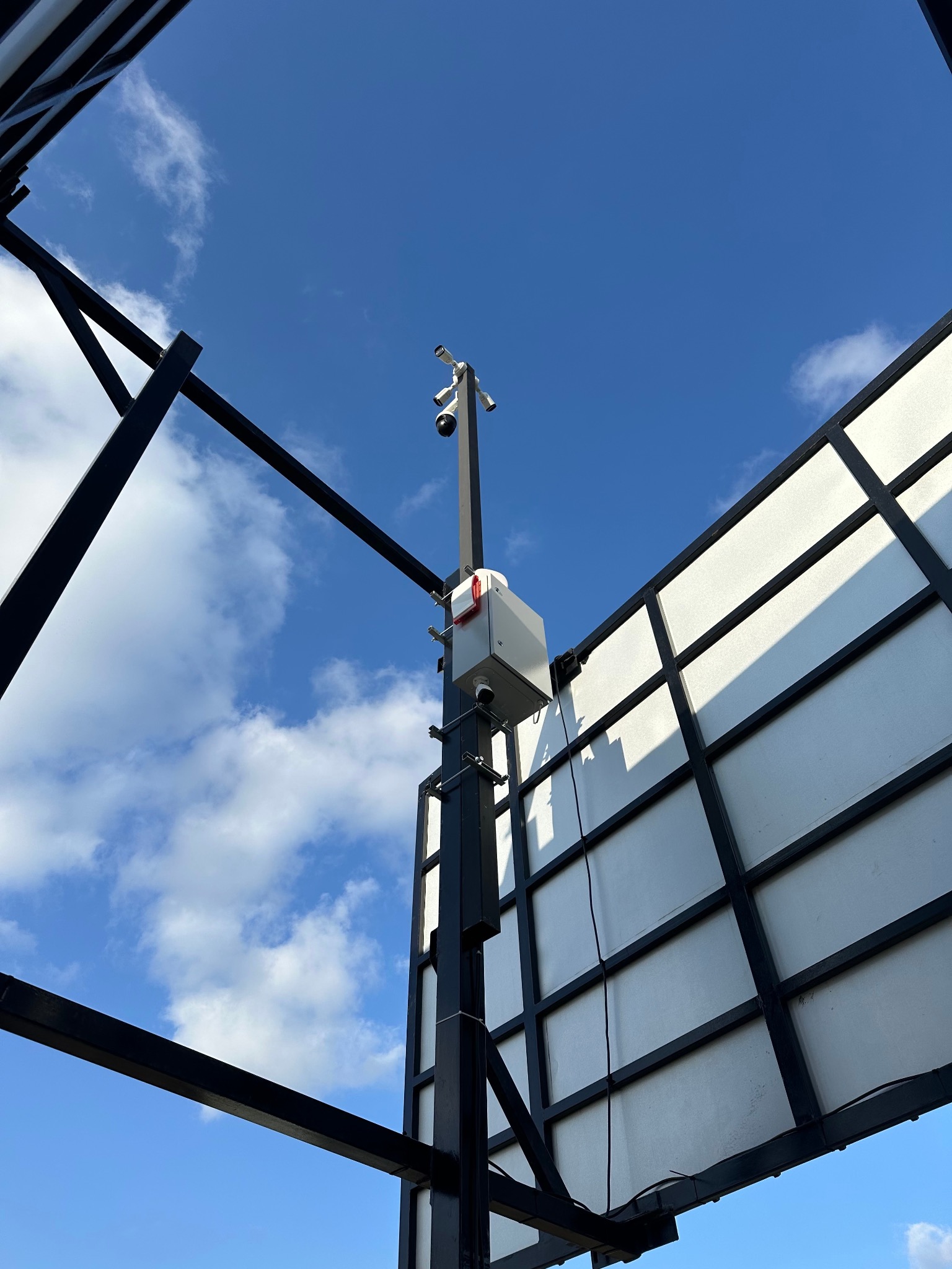 Zewnętrzny system monitoringu na konstrukcji, z widocznymi kamerami i puszką sterowniczą. Błękitne niebo z chmurami w tle. Ujęcie z dołu podkreśla wysokość instalacji.