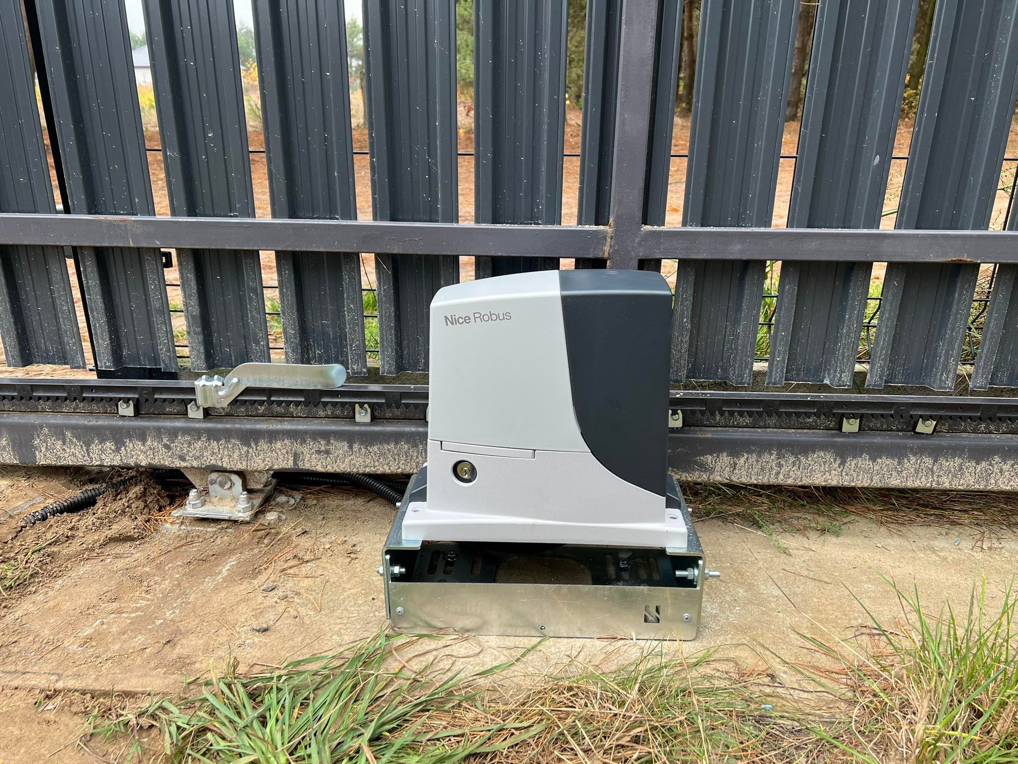 Napęd bramy przesuwnej Nice Robus zamontowany na fundamencie, z widoczną szyną bramy i elementami mocującymi. Tło stanowią drzewa i ogrodzenie panelowe.