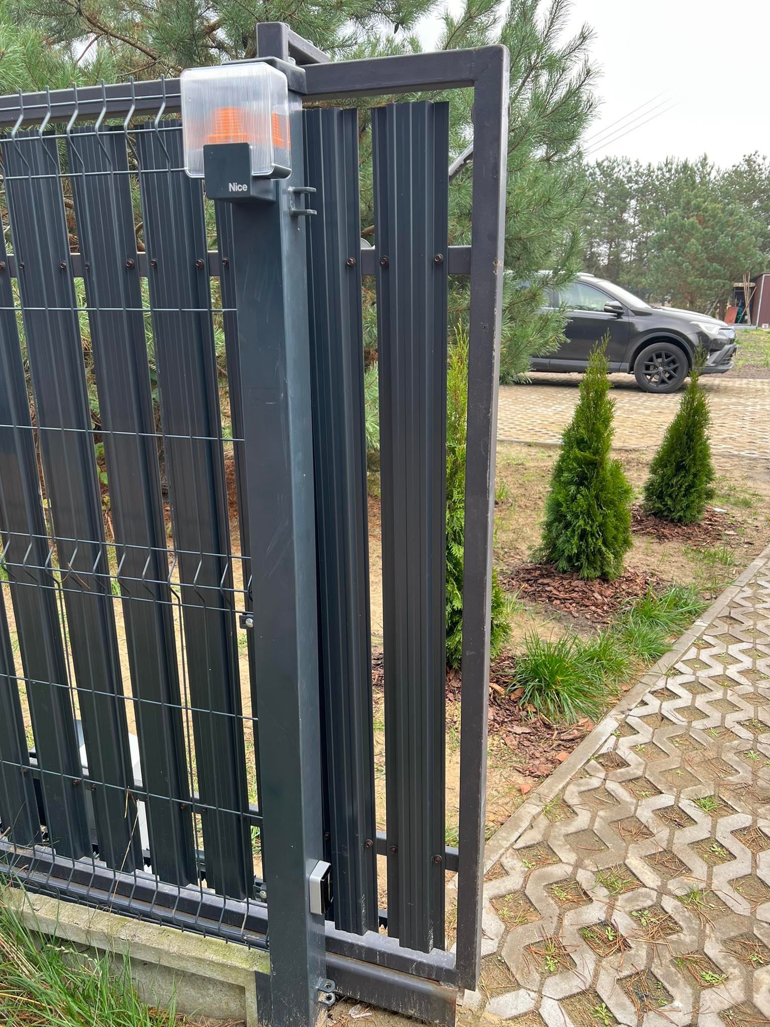 Automatyczna brama wjazdowa z lampą ostrzegawczą NICE, częściowo otwarta. W tle widoczny samochód osobowy i zieleń. Brama w kolorze grafitowym.