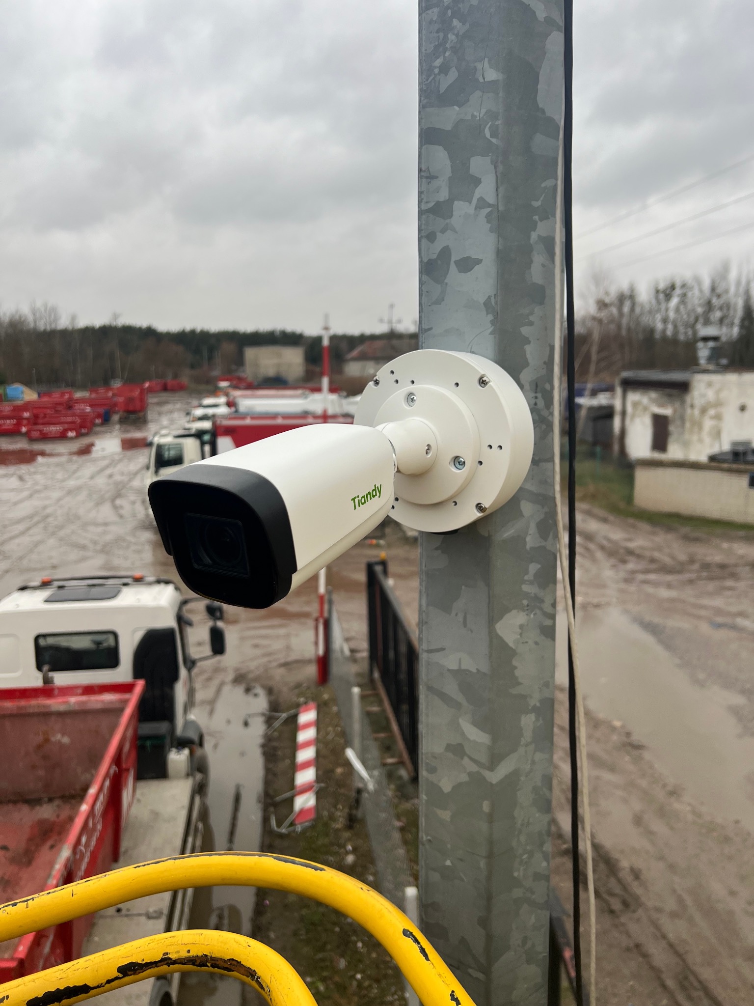 Kamera monitoringu Tiandy na metalowym słupie, z widokiem na plac budowy i ciężarówki. Szaro-biała kolorystyka, pochmurne niebo w tle. Zastosowanie monitoringu na budowie.