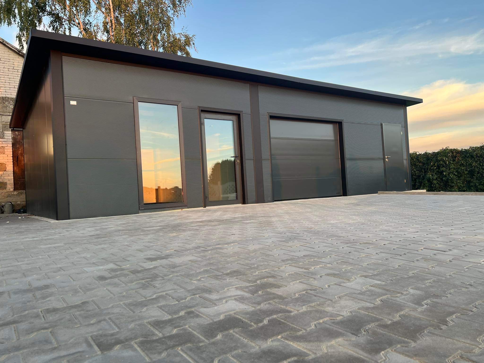 Nowoczesny, antracytowy garaż blaszany z bramą segmentową i drzwiami wejściowymi, na tle kostki brukowej i zachodzącego słońca odbijającego się w szybach.