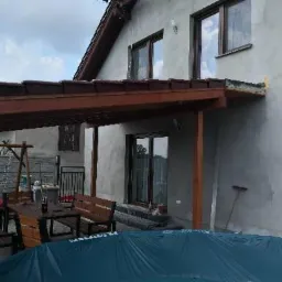 Drewniana konstrukcja z zadaszeniem przylegająca do ściany domu, widoczny stół z ławkami, huśtawka ogrodowa i przykryty basen.