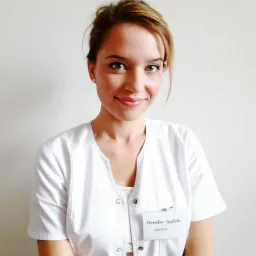 Portret uśmiechniętej kobiety w białym uniformie z identyfikatorem Monika Sajdok, na tle jasnej ściany.