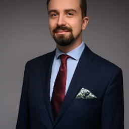 Portret uśmiechniętego mężczyzny z brodą w granatowym garniturze, bordowym krawacie i poszetką w kwiatowy wzór na szarym tle.