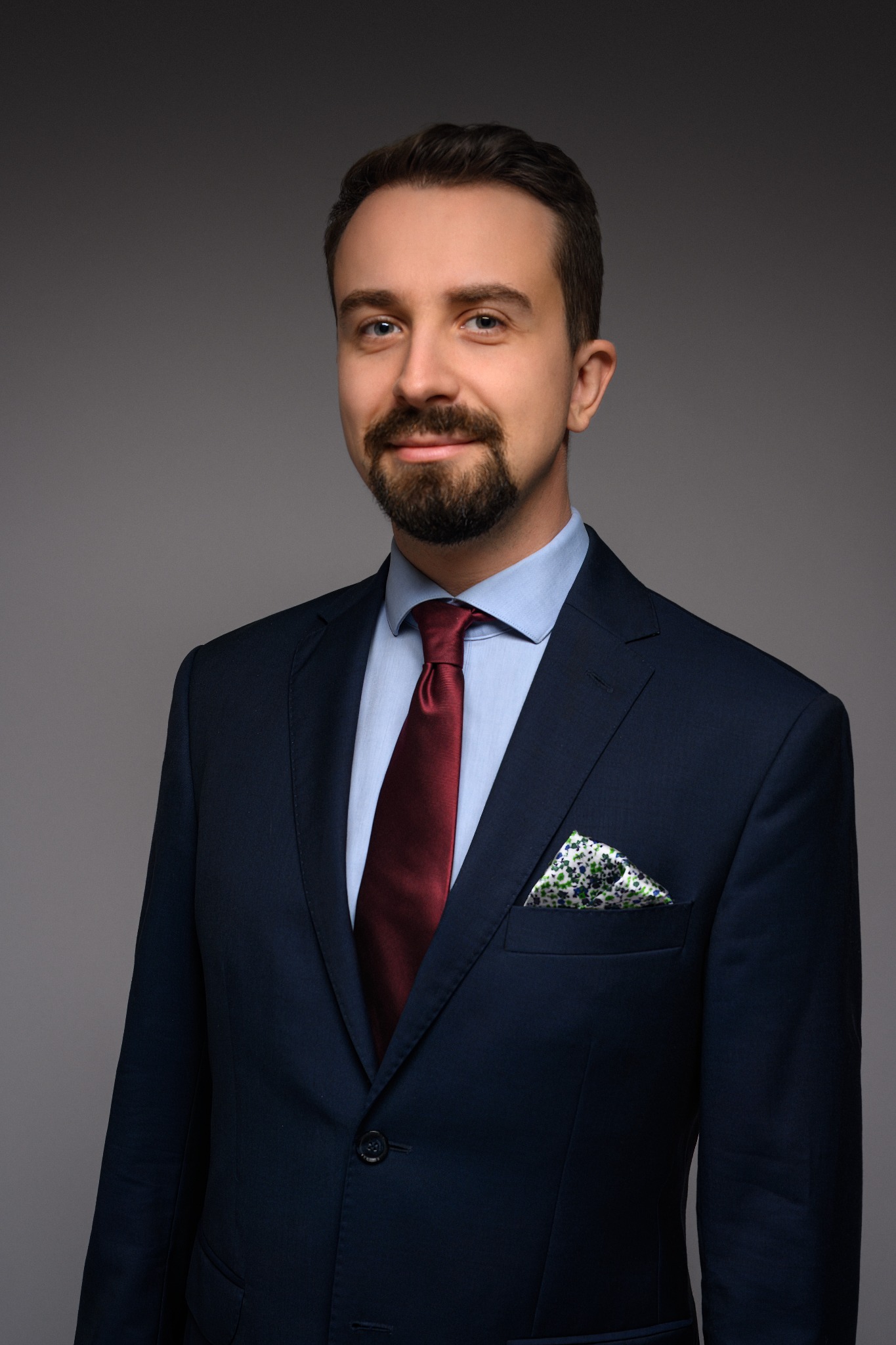 Portret uśmiechniętego mężczyzny z brodą w granatowym garniturze, bordowym krawacie i poszetką w kwiatowy wzór na szarym tle.