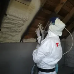 Pracownik w kombinezonie ochronnym aplikuje izolację na poddaszu, widoczne drewniane krokwie i świeżo nałożona pianka.