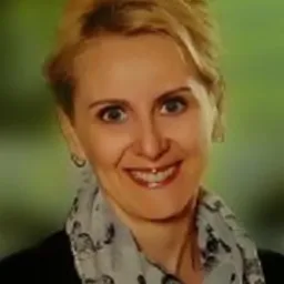 Portret uśmiechniętej kobiety z blond włosami, ubrana w czarną marynarkę i szal w wieże Eiffla, na rozmytym, zielonym tle.