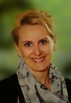 Portret uśmiechniętej kobiety z blond włosami, ubrana w czarną marynarkę i szal w wieże Eiffla, na rozmytym, zielonym tle.