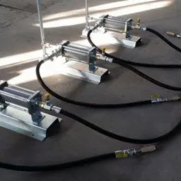 Trzy identyczne zestawy urządzeń z cylindrami pneumatycznymi na stalowych podstawach, połączone czarnymi wężami, ustawione rzędami na betonowej posadzce.