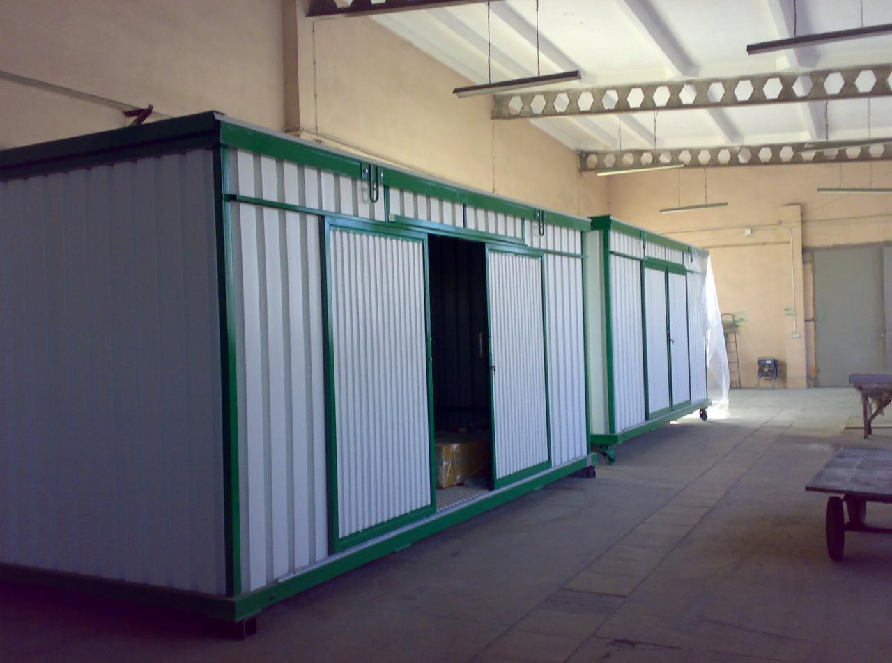 Dwa kontenery magazynowe z przesuwnymi drzwiami, pomalowane na biało z zielonymi obramowaniami, stoją w hali. Jeden kontener ma otwarte drzwi, ukazując zapakowane przedmioty w środku.