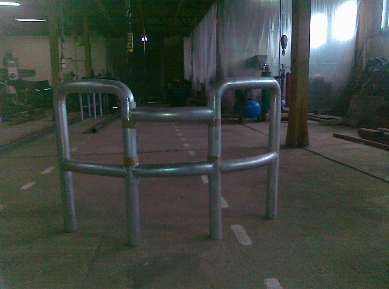 Metalowa konstrukcja ochronna, prawdopodobnie barierka, z rur spawanych na hali, z widocznymi śladami po spawaniu, na tle magazynu z niebieską butlą gazową.