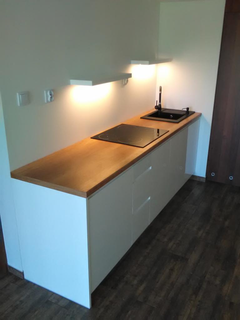 Nowoczesna zabudowa kuchenna z drewnianym blatem, białymi frontami szafek, płytą indukcyjną i czarnym zlewem, oświetlona ciepłym światłem z półek.