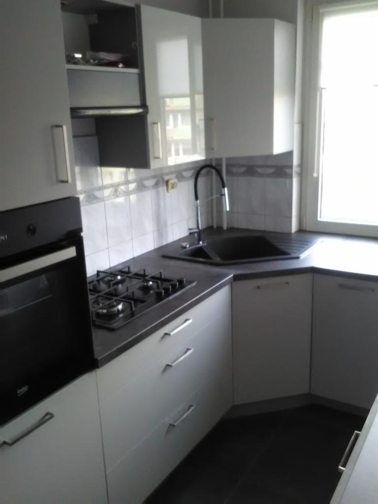 Narożna, jasna kuchnia z czarnym zlewem i płytą gazową, białe szafki z podłużnymi uchwytami, szare płytki nad blatem, widok z perspektywy.