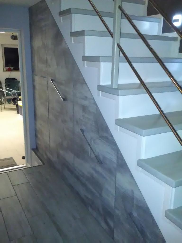 Szafa wnękowa pod schodami z szarymi frontami imitującymi beton, z metalowymi uchwytami, białe schody z metalową balustradą.