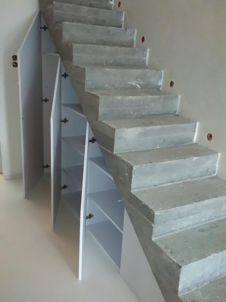 Białe szafki wnękowe pod betonowymi schodami w Goleniowie, z widocznymi zawiasami i półkami.