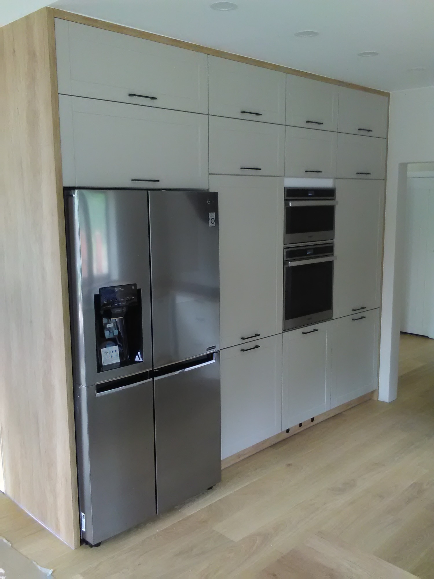 Szara kuchnia na wymiar z dużą lodówką side-by-side LG z dystrybutorem wody i kostkarką, zabudowanymi piekarnikami i szafkami górnymi z drewnianymi akcentami.