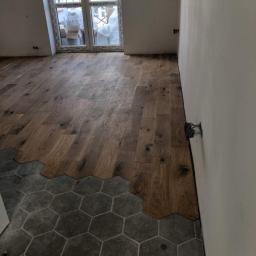 RemWag - Fragment pokoju z ułożonym parkietem w jodełkę i szarą, heksagonalną podłogą, widoczne niedokończone wykończenia przy ścianie i wystające kable.