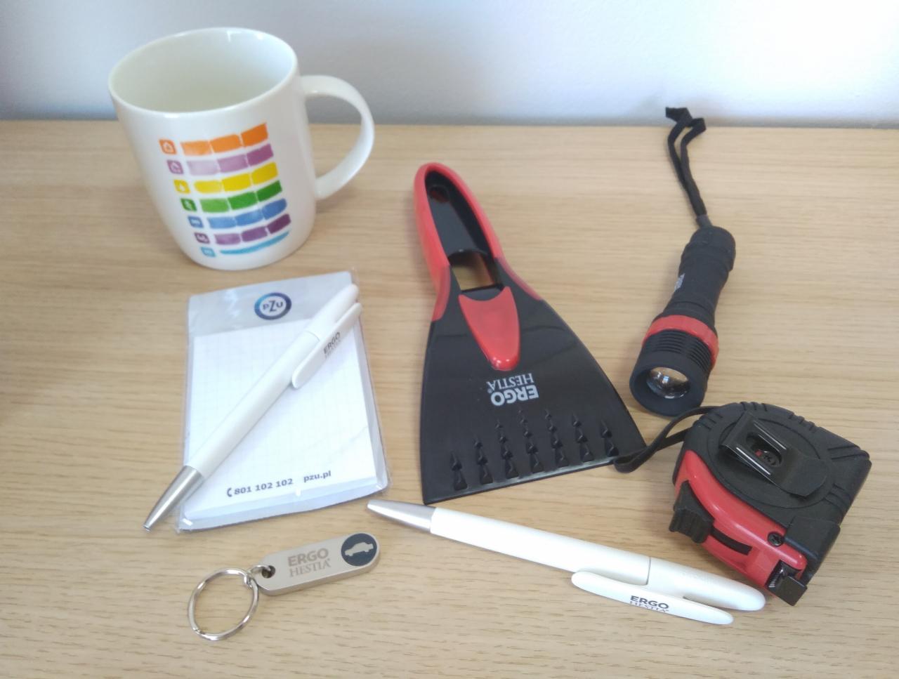 Zestaw gadżetów reklamowych: skrobaczka do szyb, miarka, latarka, długopisy, notes, breloczek i kubek z kolorowymi paskami, wszystkie z logo firmy ubezpieczeniowej.