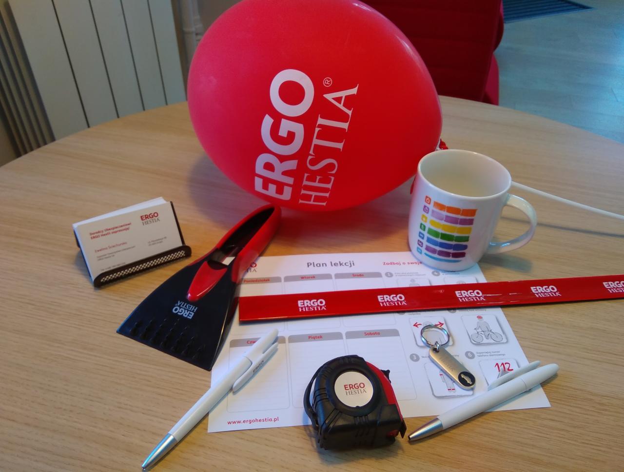 Biurko z gadżetami reklamowymi firmy ubezpieczeniowej: balon, kubek z tęczą, miarka, skrobaczka do szyb, długopisy, wizytownik, plan lekcji, smycz i brelok do kluczy.