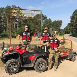 Para-Med Szkolenia Integracja - Trzech ratowników w kamizelkach odblaskowych pozuje przy czerwonym quadzie z przyczepką na tle tablicy 'Ośrodek Szkolenia Rozpoznawczego' i leśnego krajobrazu.