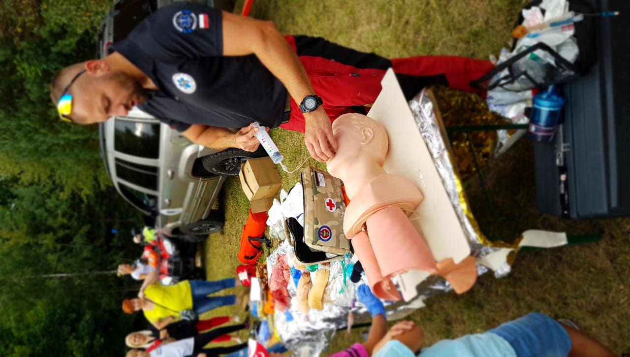 Instruktor w czarnej koszulce z naszywkami, demonstruje na manekinie podawanie leku strzykawką, w tle grupa osób obserwuje, a obok stoi samochód terenowy.