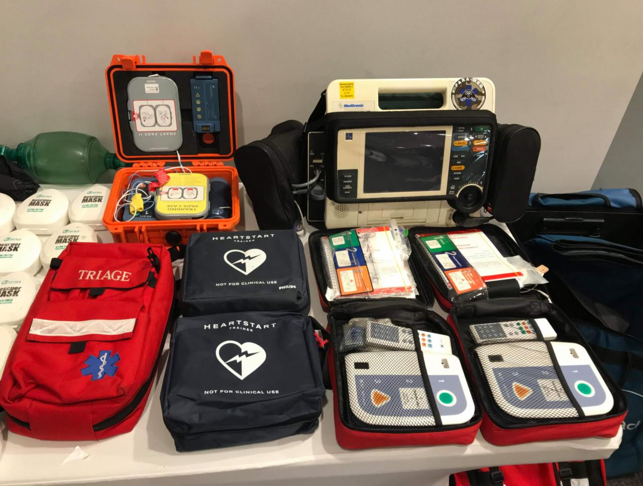 Zestaw sprzętu szkoleniowego do udzielania pierwszej pomocy: defibrylatory, maski resuscytacyjne, torba triage, elektrody treningowe, w otwartych walizkach i pokrowcach.