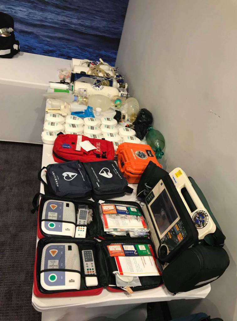 Stół z rozłożonym sprzętem medycznym: worki ambu, maski tlenowe, defibrylatory w otwartych torbach, apteczki, butle z tlenem, gotowe do użycia.
