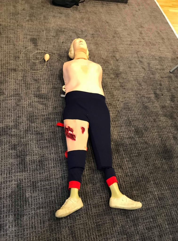 Manekin do ćwiczeń pierwszej pomocy z symulowaną raną na udzie, leżący na szarym dywanie.