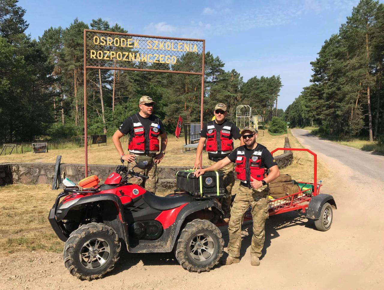 Trzech ratowników w kamizelkach odblaskowych pozuje przy czerwonym quadzie z przyczepką na tle tablicy 'Ośrodek Szkolenia Rozpoznawczego' i leśnego krajobrazu.