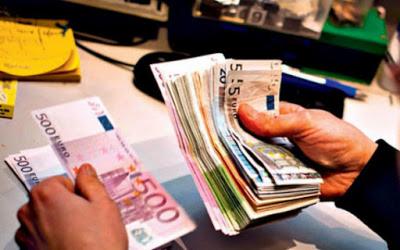 Dwie dłonie liczą plik banknotów euro o różnych nominałach, w tle biurko z akcesoriami biurowymi.