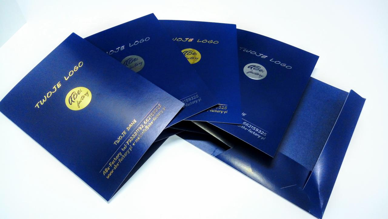 Stos niebieskich, matowych teczek firmowych z nadrukowanym złotym i srebrnym logo oraz danymi kontaktowymi na białym tle.