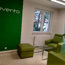 Jasne, minimalistyczne biuro z zielonym akcentem: logo 'Provento' na zielonej ścianie, skórzane zielone fotele i pufa, szklany stolik kawowy na tle okna z widokiem na drzewa.