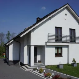 Nowoczesny dom jednorodzinny z białym tynkiem, ciemną stolarką okienną i balkonem z metalowymi barierkami. Podjazd wyłożony kostką brukową, z nasadzeniami roślin ozdobnych.
