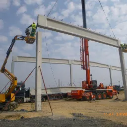 Montaż prefabrykowanych elementów konstrukcyjnych hali z użyciem dźwigu i podnośników koszowych, widoczne stalowe podpory tymczasowe.
