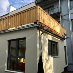 Rozbudowa domu z drewnianą balustradą na dachu, widok z dołu, z czarną kurtką wiszącą na rynnie.