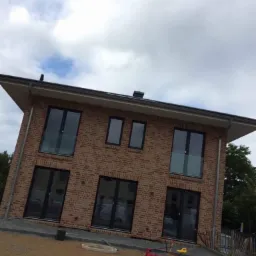 Dwukondygnacyjny budynek z czerwonej cegły z czarnymi ramami okiennymi i balkonami, widoczny z perspektywy placu budowy z zabawkami dla dzieci i taczką na pierwszym planie.