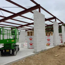 Wznoszenie ścian z betonu komórkowego na fundamencie z wylewką, widoczna konstrukcja stalowa dachu i podnośnik nożycowy.