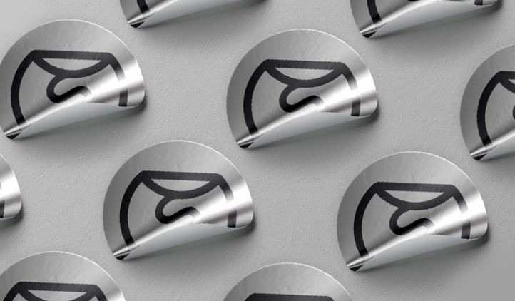 Srebrne, okrągłe etykiety z czarnym, minimalistycznym logo na szarym tle. Częściowo odklejone, ukazujące błyszczący spód.