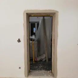 Przygotowanie do montażu ościeżnicy drzwiowej: szpachlowanie i wyrównywanie ścian wokół otworu drzwiowego, widoczna poziomica i worek ze szpachlą w tle.