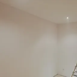 Pomalowane na biało pomieszczenie z aluminiową drabiną opartą o ścianę, widoczny punktowy reflektor w suficie.