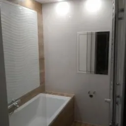 Nowoczesna łazienka z białą wanną obudowaną panelami imitującymi drewno, dekoracyjną białą płytką ścienną z falistym wzorem, lustrem w srebrnej ramie i grzejnikiem drabinkowym. Oświetlenie punktowe...